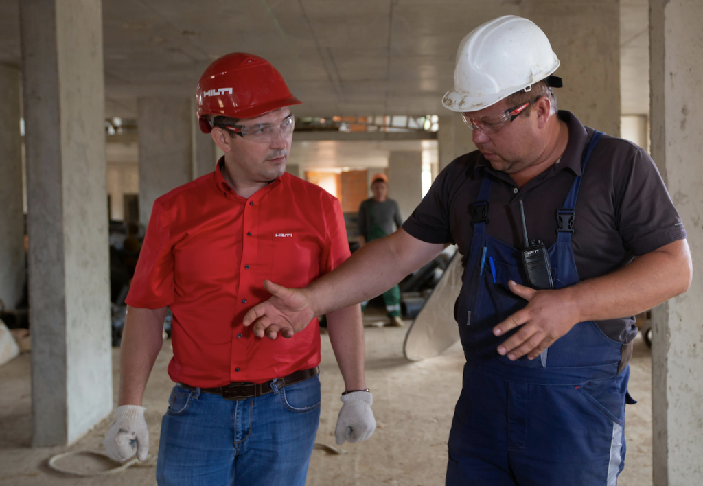 Das Foto zeigt zwei Männer mit Schutzhelm im Inneren eines im Bau befindlichen Gebäudes, wobei der eine Mann dem anderen Mann etwas zeigt und erläutert.