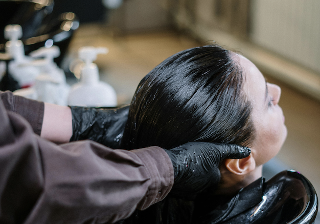 Das Foto zeigt eine Kundin, der vom Friseur am Waschtisch die Haare gewaschen werden. Der Friseur trägt dünne schwarze Handschuhe, während er mit Shampoo und Wasser hantiert.