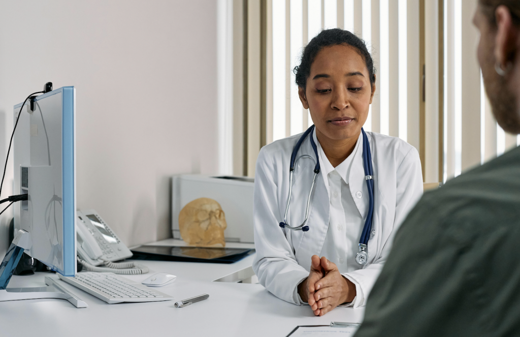 Das Foto zeigt eine Ärztin an ihrem Schreibtisch während der Beratung eines Klienten. Dabei blickt sie auf ein Dokument, das auf dem Tisch liegt.