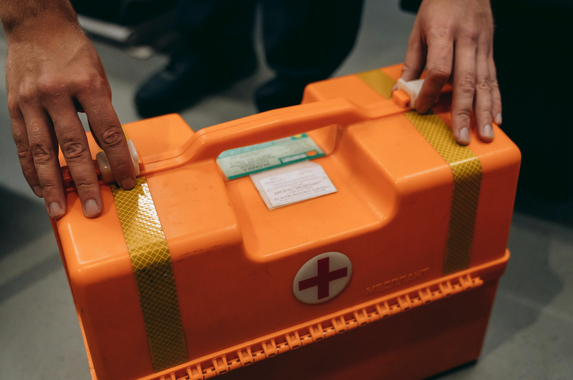 Das Foto zeigt einen Erste-Hilfe-Koffer auf dem Boden stehend, dahinter hockt eine Person, deren zwei Hände den Verschluss des Kastens öffnen wird.